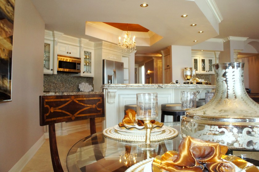 Here you can see the dramatic change in the kitchen! In the upper cabinets, the glass shelves and mirrored backs help open them up and add depth.  The strange placement of gold paint on the inside of the ceiling tray was replaced with an extension of the new ceiling color.  The crown was painted off-white, and I added a gold shimmering grasscloth to the ceiling, inside the recessed tray.  This provided drama and emphasis to this ceiling treatment, but in a more natural, aesthetically-pleasing way. 